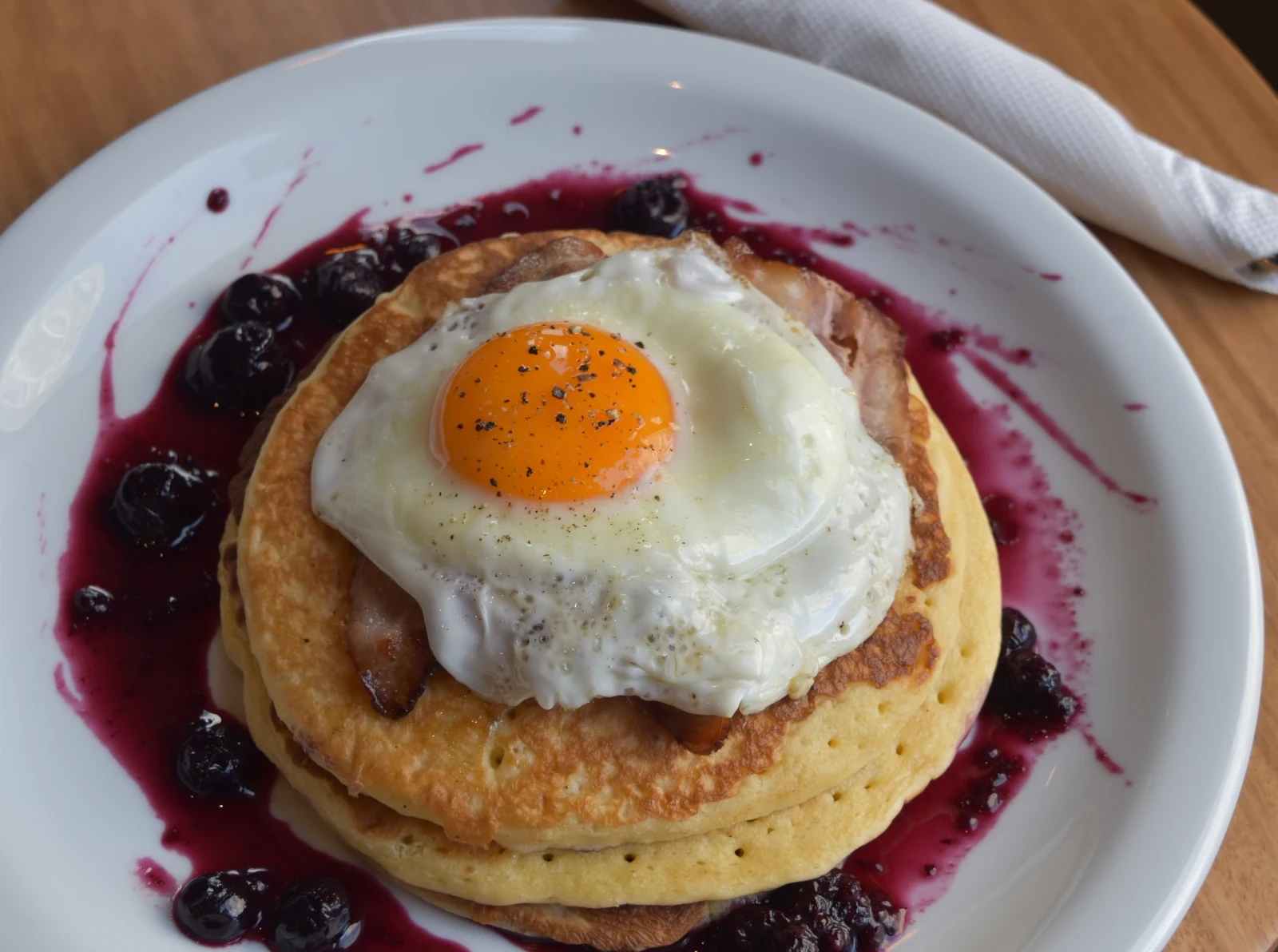 Primer plano de un plato de hot cakes americanos de Café Tapiz, cubiertos con un huevo frito, tocino y una salsa de frutos del bosque